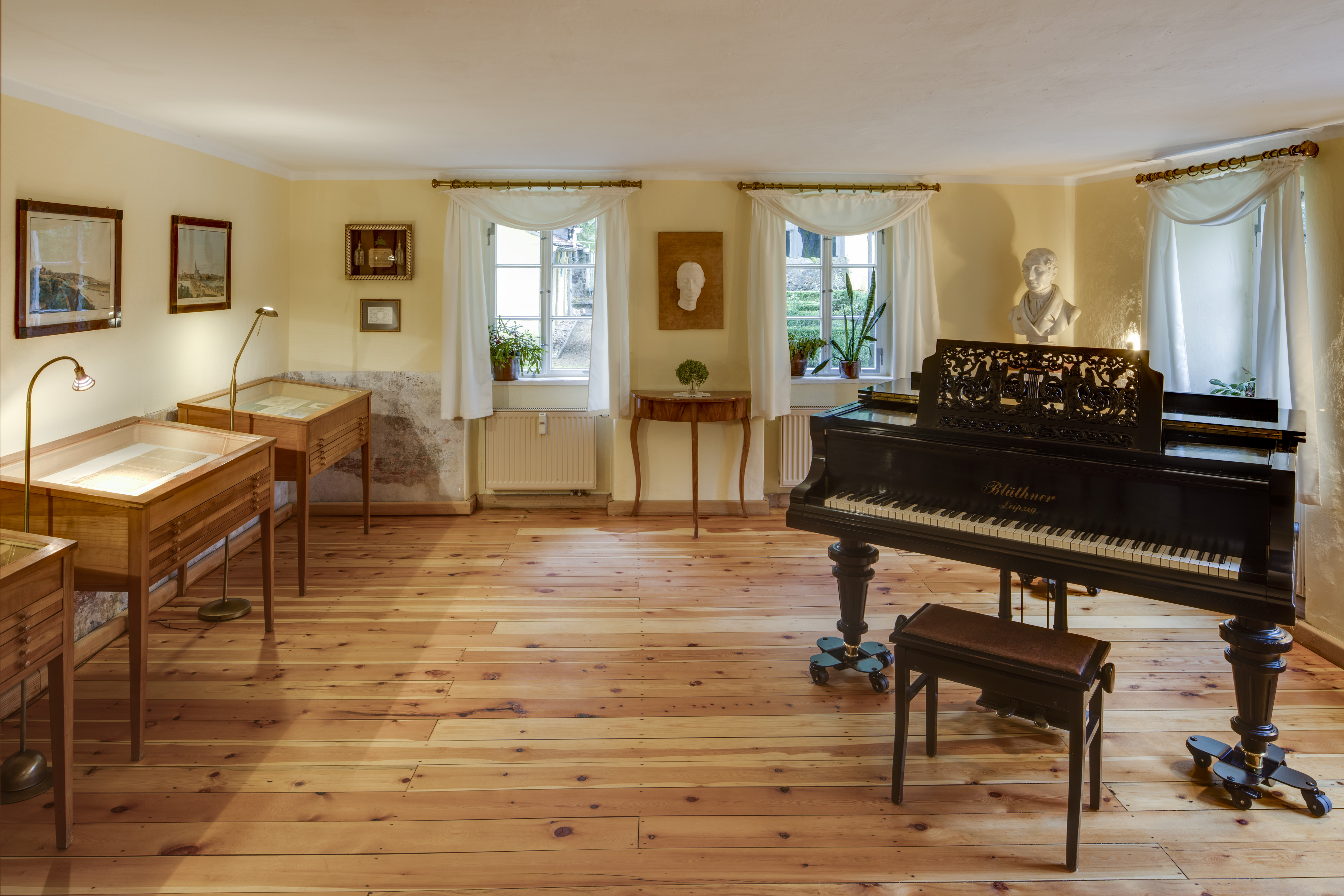 KI generiert: Das Bild zeigt einen gepflegten Raum mit Holzboden, der als Museum eingerichtet ist. Im Vordergrund befindet sich ein schwarzes Klavier, und entlang der Wände stehen Vitrinen mit Ausstellungsstücken sowie eine Büste auf einem Sockel.