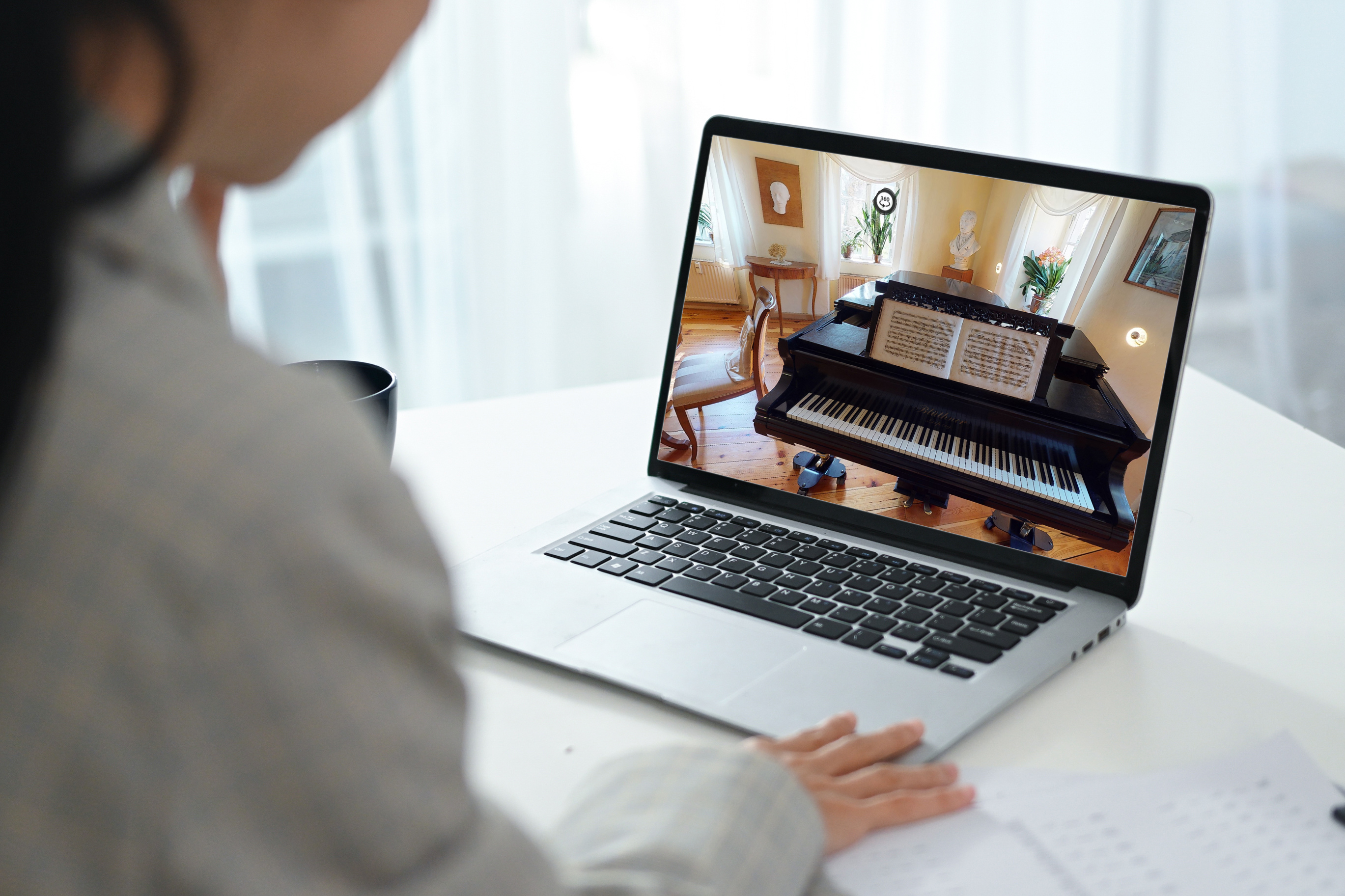 KI generiert: Eine Person betrachtet auf einem Laptop das Bild eines Flügels in einem elegant eingerichteten Raum. Im Vordergrund liegt ein Notenblatt auf dem Tisch neben dem Laptop.