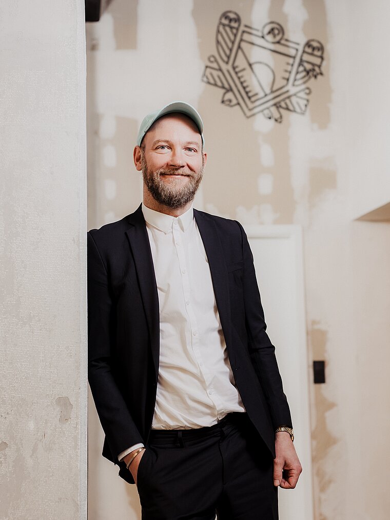 AI-generated: A man in a suit is leaning casually against a wall, wearing a baseball cap and smiling. The room has an industrial style, with a graphic element on the wall in the background.