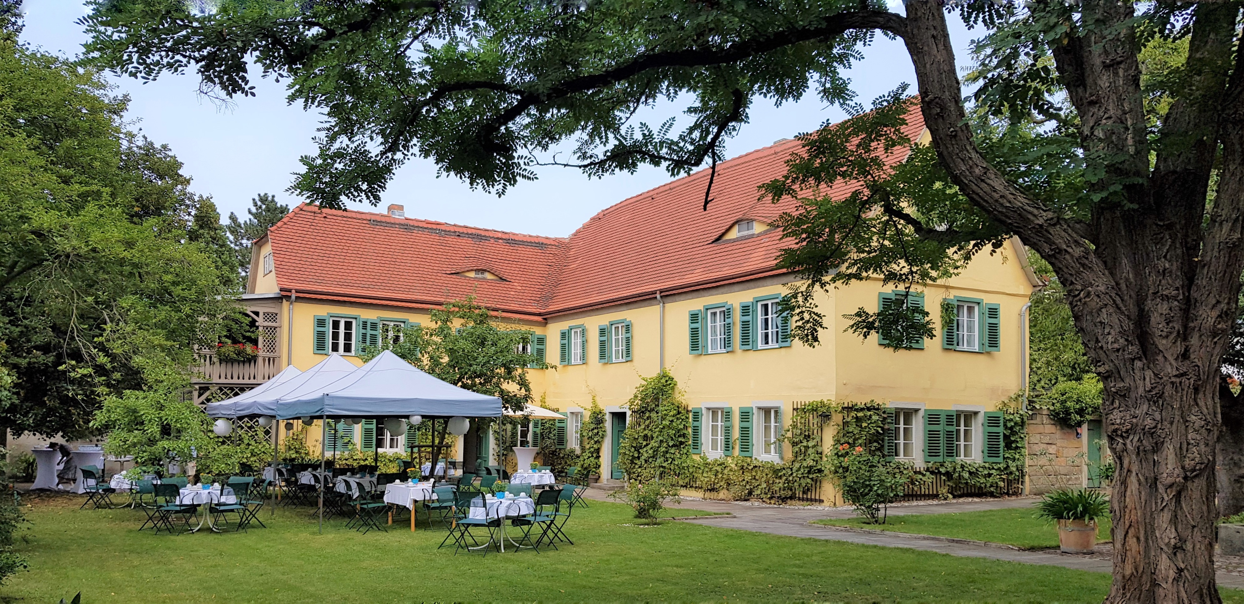 KI generiert: Das Bild zeigt ein großes, gelbes Gebäude mit roten Dächern und grünen Fensterläden, umgeben von einem gepflegten Garten mit Bäumen. Im Vordergrund sind mehrere Tische und Stühle unter weißen Pavillons aufgestellt.