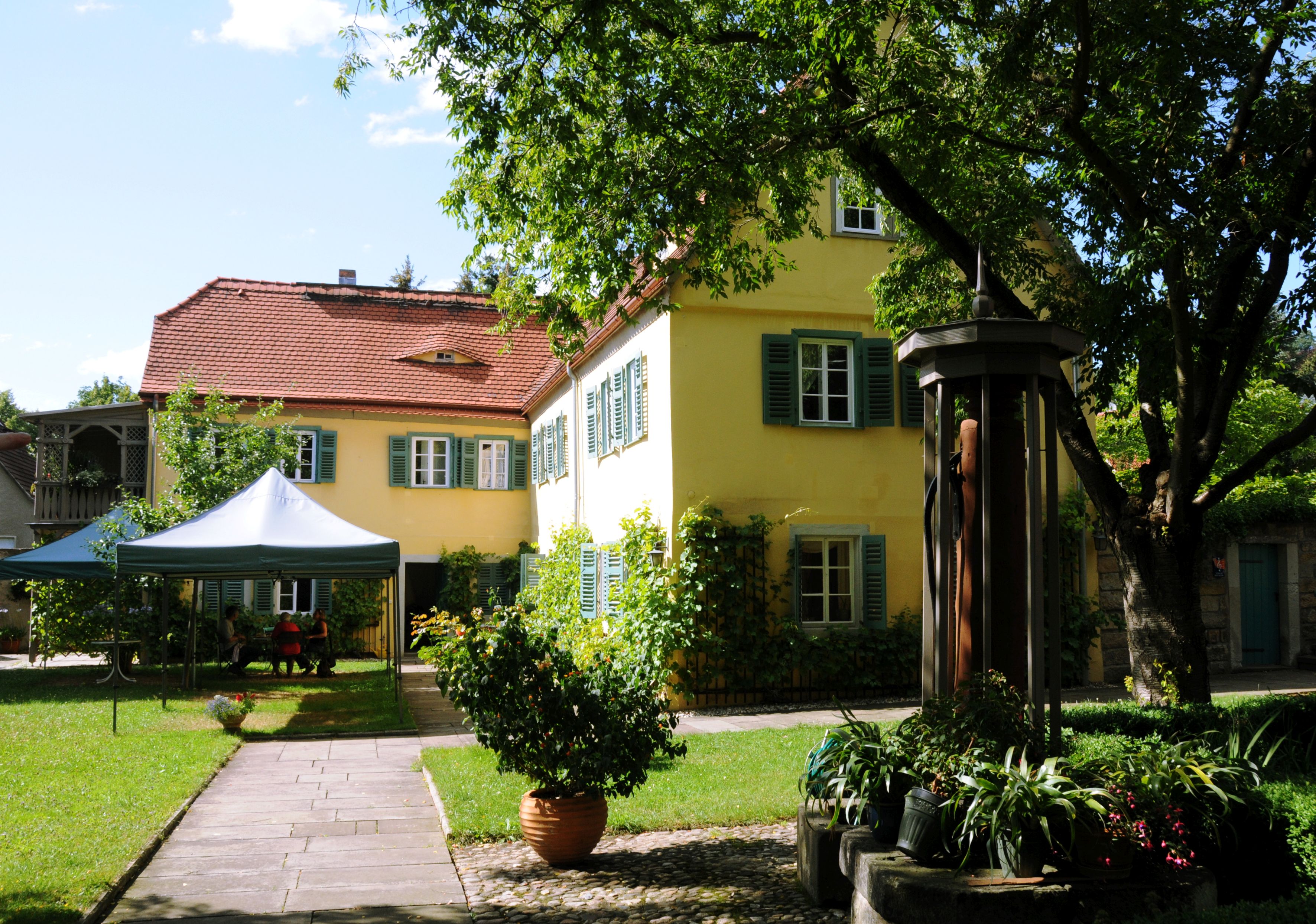 KI generiert: Das Bild zeigt ein malerisches, zweistöckiges Haus mit gelben Wänden und roten Dächern, umgeben von üppiger Vegetation. Im Vordergrund befindet sich ein gepflegter Garten mit einem begehbaren Weg, einem Pavillon und verschiedenen Pflanzen.