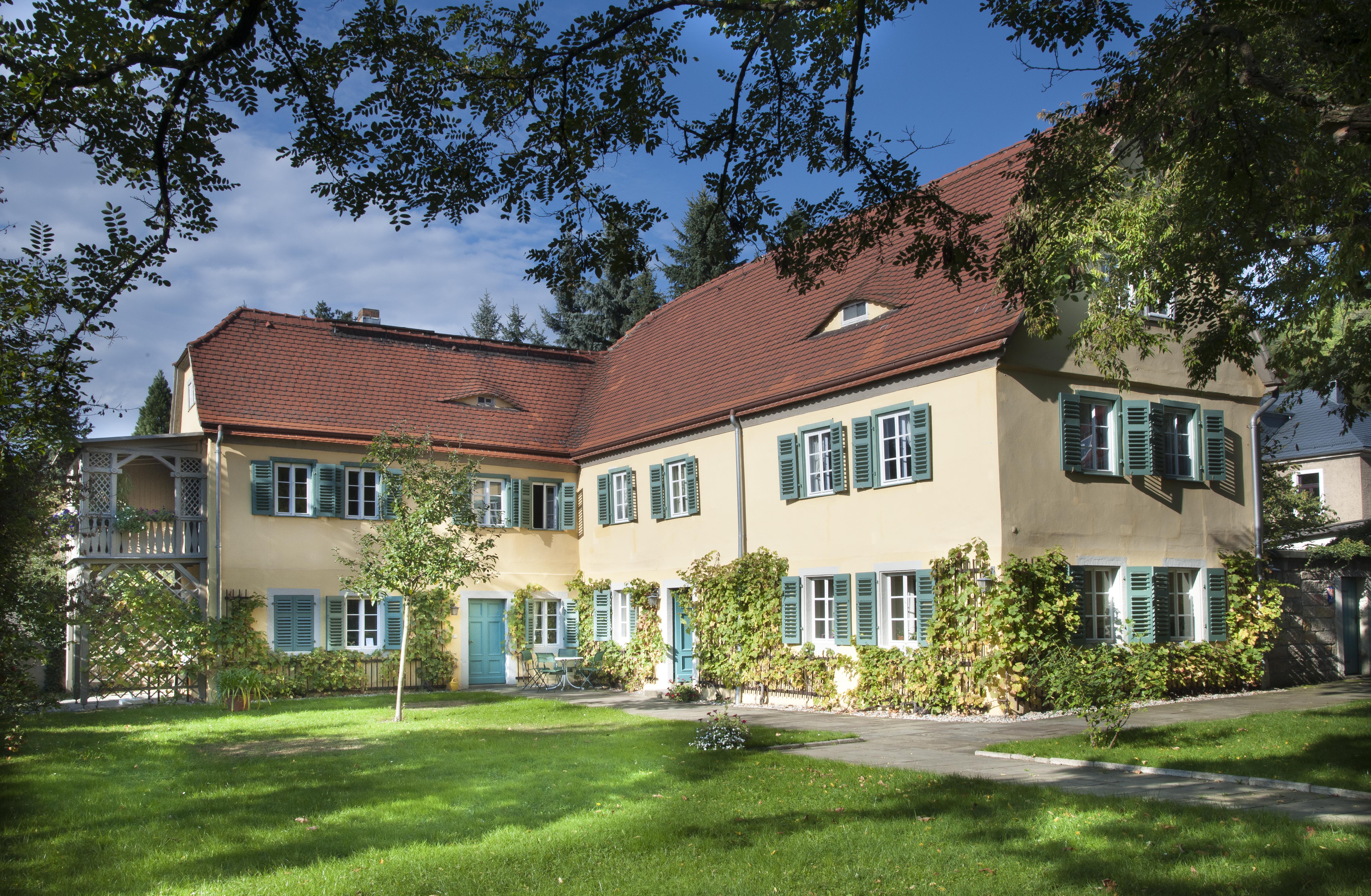 AI generated: The image shows a large, traditional building with yellow walls and blue shutters, surrounded by a green garden. The sky is clear and the trees frame the house harmoniously.